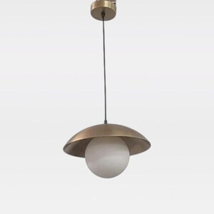 Könnte beinhalten: Eine Pendelleuchte mit einem kreisförmigen, messingfarbenen Metallschirm in Form einer breiten, flachen Kuppel. Eine kugelförmige weiße Glaskugel hängt mittig unter dem Schirm und ist über ein Kabel mit einem passenden Deckenbaldachin verbunden.