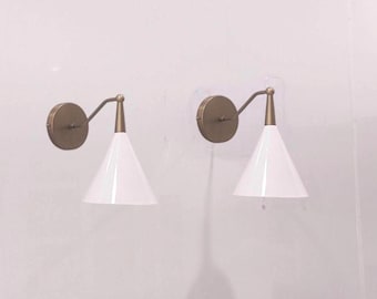 May include: A pair of wall sconces featuring circular brass-toned backplates, angled arms, and white conical shades. The fixtures are mounted to a wall, showcasing a clean design with metallic hardware and smooth, white, downward-facing lamp shades.