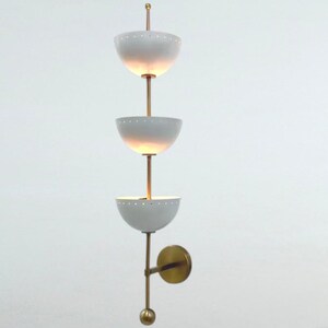 May include: A wall-mounted light fixture featuring a vertical brass rod supporting three white, bowl-shaped shades. Each shade has a row of small perforations along the upper rim. The fixture includes a circular brass wall plate and a spherical finial.