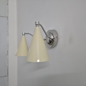 Puede incluir: Aplique de pared con dos pantallas c&oacute;nicas de color crema. La l&aacute;mpara tiene un acabado cromado pulido y est&aacute; montada en una pared blanca. El dise&ntilde;o es sencillo y moderno, adecuado para varios estilos de interior.