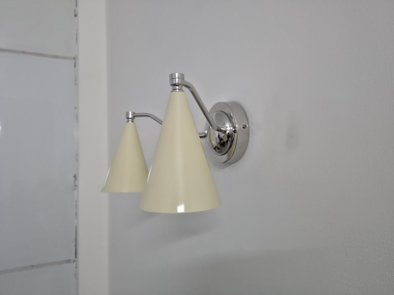 Puede incluir: Aplique de pared con dos pantallas c&oacute;nicas de color crema. La l&aacute;mpara tiene un acabado cromado pulido y est&aacute; montada en una pared blanca. El dise&ntilde;o es sencillo y moderno, adecuado para varios estilos de interior.