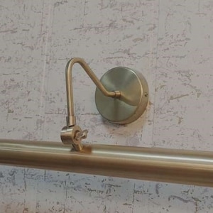 May include: A brass-toned picture light fixture. The fixture has a long, cylindrical light bar, a curved arm, and a circular wall mount. The light is designed to illuminate artwork or other wall-mounted items.
