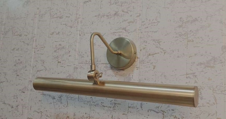 May include: A brass-toned picture light fixture. The fixture has a long, cylindrical light bar, a curved arm, and a circular wall mount. The light is designed to illuminate artwork or other wall-mounted items.