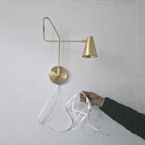 Op de afbeelding: Een messingkleurige wandlamp met een conische kap. De lamp heeft een ronde basis en een verstelbare arm. Een wit snoer loopt van de lamp, vastgehouden door een persoon.