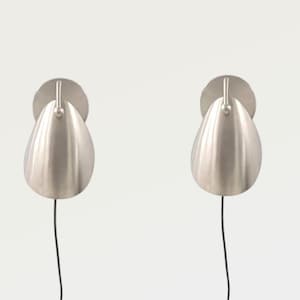 Könnte beinhalten: Ein Paar Wandleuchten mit gebürstetem Silber-Finish. Jede Lampe hat einen abgerundeten, tropfenförmigen Metallschirm, der mit einem kleinen vertikalen Arm an einer runden Wandplatte befestigt ist. Ein dünnes schwarzes Kabel hängt von jeder Leuchte.
