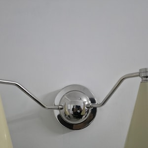Puede incluir: L&aacute;mpara de pared con dos pantallas c&oacute;nicas de color crema. La l&aacute;mpara tiene un acabado cromado pulido, incluyendo la base circular y los brazos curvos que sostienen las pantallas. Dise&ntilde;o moderno y minimalista.
