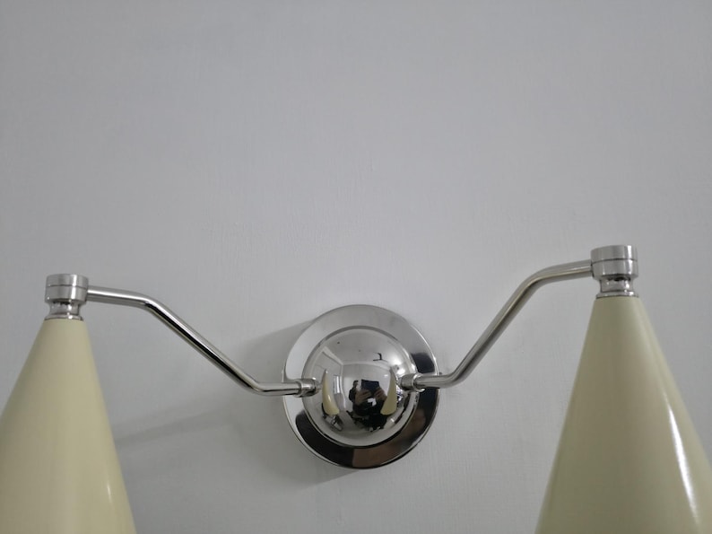 Puede incluir: L&aacute;mpara de pared con dos pantallas c&oacute;nicas de color crema. La l&aacute;mpara tiene un acabado cromado pulido, incluyendo la base circular y los brazos curvos que sostienen las pantallas. Dise&ntilde;o moderno y minimalista.