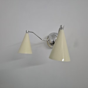 Puede incluir: Una l&aacute;mpara de pared con acabado cromado pulido y dos pantallas en forma de cono de color crema. La l&aacute;mpara tiene un brazo curvo que conecta las pantallas a una base circular.
