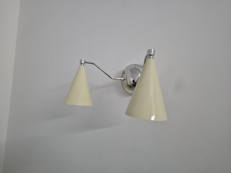 Puede incluir: Una l&aacute;mpara de pared con acabado cromado pulido y dos pantallas en forma de cono de color crema. La l&aacute;mpara tiene un brazo curvo que conecta las pantallas a una base circular.