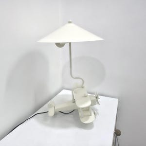 Könnte beinhalten: Eine skurrile, cremefarbene Tischlampe in Flugzeugform. Die Lampe hat ein Doppeldecker-Design mit Propeller und konischem Lampenschirm. Die Lampe steht auf einer weißen Oberfläche, mit einem schwarzen Netzkabel.