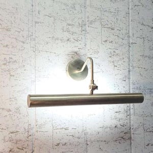 May include: A brass-toned picture light fixture with a cylindrical light bar and a curved arm. The light is mounted on a circular base and is positioned against a textured, light-colored wall.