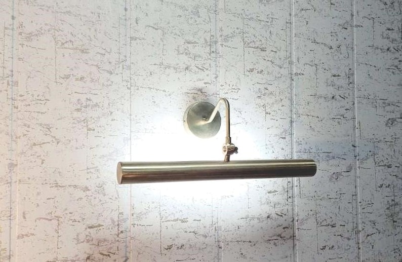 May include: A brass-toned picture light fixture with a cylindrical light bar and a curved arm. The light is mounted on a circular base and is positioned against a textured, light-colored wall.