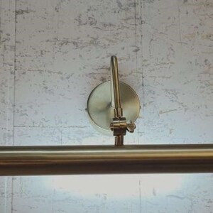 May include: A brass-toned picture light fixture with a cylindrical light bar and a circular wall mount. The light bar is positioned horizontally, and the fixture is mounted on a textured, light-colored wall.