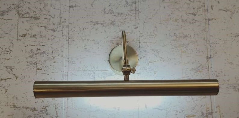 May include: A brass-toned picture light fixture with a cylindrical light bar and a circular wall mount. The light bar is positioned horizontally, and the fixture is mounted on a textured, light-colored wall.