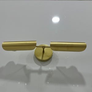 Pode incluir: Uma arandela de parede dourada com duas c&uacute;pulas cil&iacute;ndricas. A arandela tem um design simples e moderno.