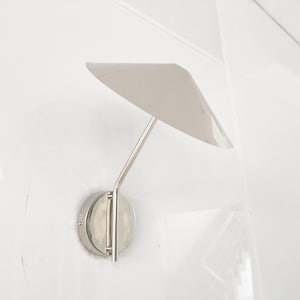 Op de afbeelding: Een moderne, zilverkleurige wandlamp met een minimalistisch ontwerp. Het heeft een cirkelvormige basis, een slanke arm en een driehoekige kap. De armatuur is op een witte muur gemonteerd, wat de strakke, eigentijdse esthetiek benadrukt.