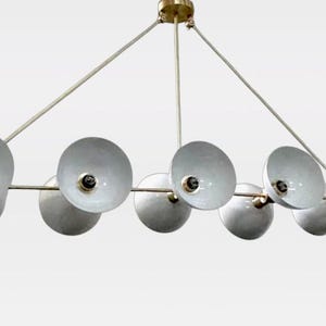 Könnte beinhalten: Ein linearer Kronleuchter mit sechs weißen, schüsselförmigen Metallschirmen, die horizontal angeordnet sind. Die Leuchte wird von einem dünnen, goldfarbenen Metallrahmen mit einem zentralen Befestigungspunkt und abgewinkelten Armen gehalten.