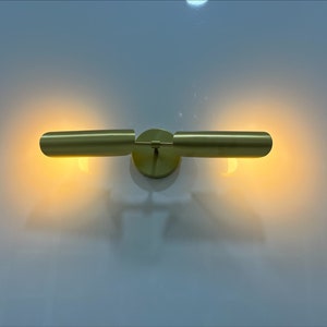 Pode incluir: Uma arandela de parede dourada com duas lumin&aacute;rias cil&iacute;ndricas. As luzes est&atilde;o ligadas e projetam um brilho quente.