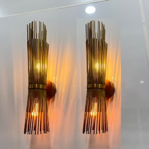 Könnte beinhalten: Zwei goldene Wandleuchten mit einem einzigartigen Design aus dünnen, vertikalen Metallstäben. Die Leuchten sind beleuchtet und strahlen ein warmes Licht aus.