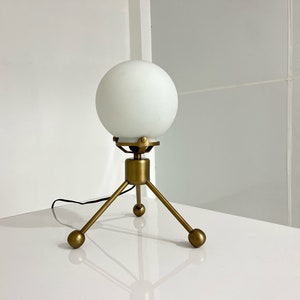Könnte beinhalten: Eine weiße, mattierte Glaskugellampe mit einem goldenen Metall-Dreibeinständer. Die Lampe hat ein modernes Design und ist perfekt für einen Nachttisch oder Schreibtisch.