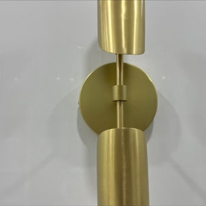 Pode incluir: Uma arandela de parede dourada com duas lumin&aacute;rias cil&iacute;ndricas. A arandela tem uma base redonda e um bra&ccedil;o girat&oacute;rio que permite ajustar as luzes.