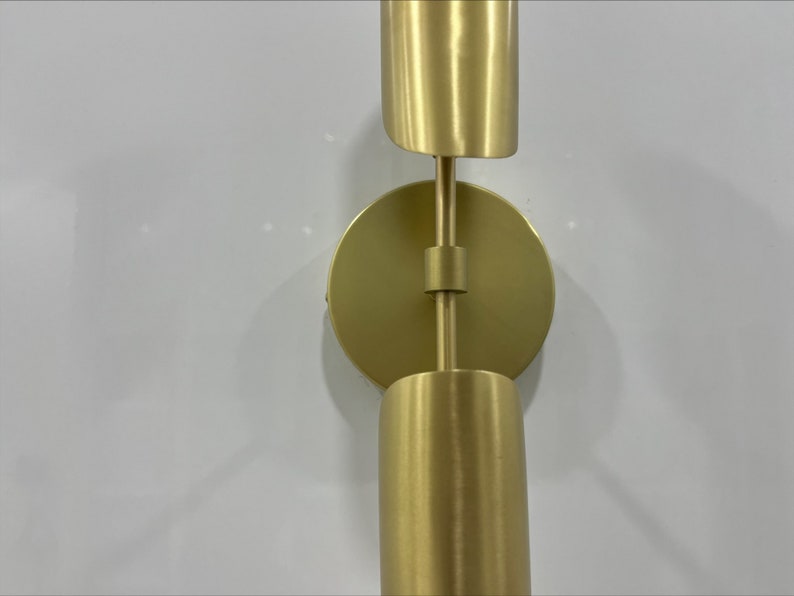Pode incluir: Uma arandela de parede dourada com duas lumin&aacute;rias cil&iacute;ndricas. A arandela tem uma base redonda e um bra&ccedil;o girat&oacute;rio que permite ajustar as luzes.