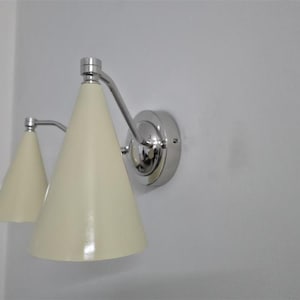 Puede incluir: Una l&aacute;mpara de pared con dos pantallas c&oacute;nicas de color crema. La l&aacute;mpara tiene un acabado cromado pulido y est&aacute; montada en una pared blanca. El dise&ntilde;o es moderno y minimalista.