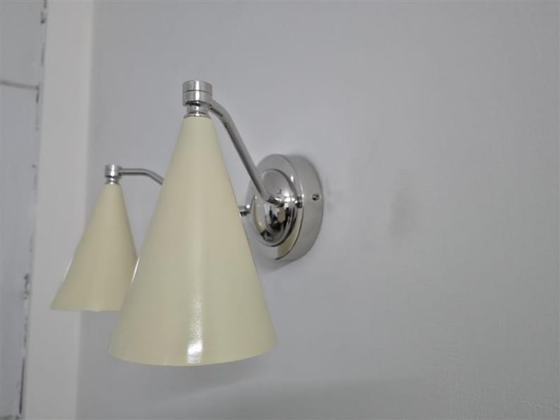 Puede incluir: Una l&aacute;mpara de pared con dos pantallas c&oacute;nicas de color crema. La l&aacute;mpara tiene un acabado cromado pulido y est&aacute; montada en una pared blanca. El dise&ntilde;o es moderno y minimalista.