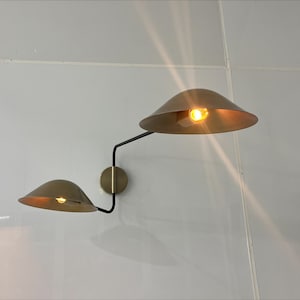 May include: A modern wall sconce with two gold-toned shades and a black metal arm. The shades are angled in opposite directions, creating a unique and stylish look.