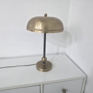 Könnte beinhalten: Eine messingfarbene Schreibtischlampe mit einem gewölbten Schirm und einem schwarzen Stiel. Die Lampe hat einen runden Sockel und einen kleinen Knopf oben. Die Lampe steht auf einer weißen Oberfläche.