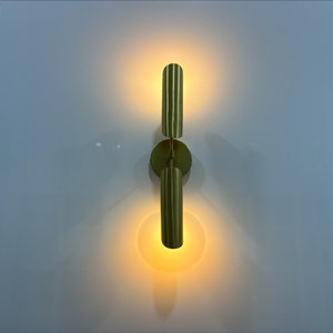 Pode incluir: Uma arandela de parede dourada com duas lumin&aacute;rias verticais. As lumin&aacute;rias est&atilde;o ligadas e emitem um brilho quente.
