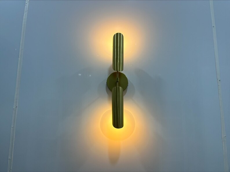 Pode incluir: Uma arandela de parede dourada com duas lumin&aacute;rias verticais. As lumin&aacute;rias est&atilde;o ligadas e emitem um brilho quente.