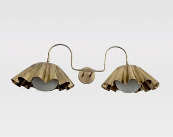 May include: A double wall sconce featuring two brass-toned, ruffled, bell-shaped shades. Each shade houses a spherical, frosted white glass bulb. The fixtures are connected by curved metal arms extending from a central circular wall mount.