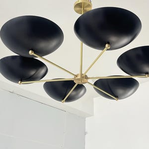 May include: A contemporary chandelier featuring six black, bowl-shaped shades. The shades are suspended from brass-coloured arms and a central fitting. The fitting is mounted on a white ceiling, creating a striking contrast.