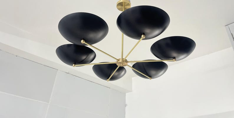 May include: A contemporary chandelier featuring six black, bowl-shaped shades. The shades are suspended from brass-coloured arms and a central fitting. The fitting is mounted on a white ceiling, creating a striking contrast.