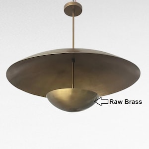 Könnte beinhalten: Eine moderne Pendelleuchte aus Rohmessing. Sie hat einen großen, flachen, scheibenförmigen Schirm über einem kleineren, schalenförmigen Schirm. Die Leuchte hängt an einer einzelnen Stange und einer kreisförmigen Deckenhalterung.