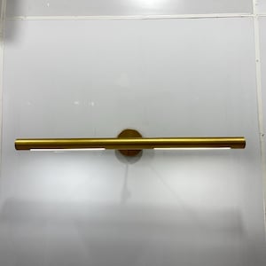 Könnte beinhalten: Eine goldfarbene Wandleuchte mit einem langen, dünnen, rechteckigen Leuchtbalken. Der Leuchtbalken ist an einer runden, kreisförmigen Basis befestigt.