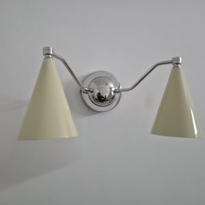 Puede incluir: Una l&aacute;mpara de pared con acabado cromado pulido. Cuenta con dos pantallas c&oacute;nicas de color crema. La l&aacute;mpara est&aacute; montada en una pared blanca, ofreciendo una est&eacute;tica limpia y moderna. Las pantallas miden unos 15 cm de alto.
