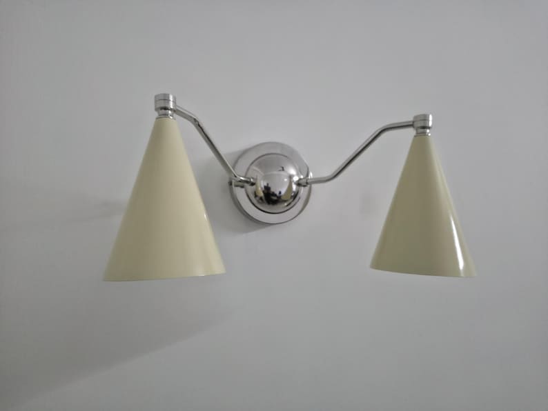 Puede incluir: Una l&aacute;mpara de pared con acabado cromado pulido. Cuenta con dos pantallas c&oacute;nicas de color crema. La l&aacute;mpara est&aacute; montada en una pared blanca, ofreciendo una est&eacute;tica limpia y moderna. Las pantallas miden unos 15 cm de alto.