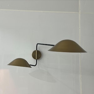May include: A wall-mounted light fixture with two gold-colored shades and a black metal arm. The shades are angled downwards and the arm is curved.