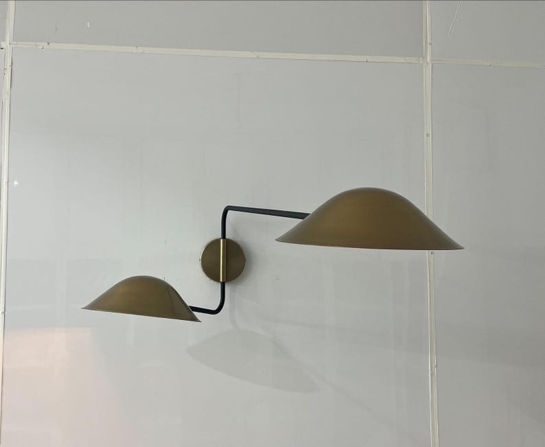 May include: A wall-mounted light fixture with two gold-colored shades and a black metal arm. The shades are angled downwards and the arm is curved.