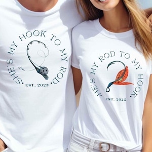 Może przedstawiać: Dwie białe koszulki z nadrukiem w temacie wędkowania. Pierwsza koszulka ma wędkę i napis "She's my hook to my rod. Est. 2025". Druga koszulka ma haczyk i napis "He's my rod to my hook. Est. 2025".