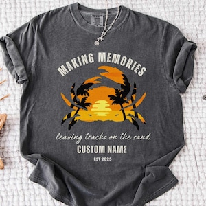 Puede incluir: Una camiseta gris oscuro con un gráfico de una silueta de cangrejo con una puesta de sol y palmeras. El texto "Making Memories leaving tracks on the sand CUSTOM NAME EST 2025" está impreso en la camiseta.