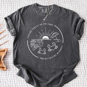 Puede incluir: Una camiseta gris oscuro con un diseño gráfico blanco de una escena de playa con un sol, olas, una sombrilla y dos cangrejos. El texto en el gráfico dice "Leaving tracks on the sand making lasting memories".