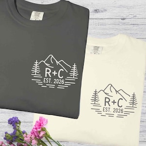 Op de afbeelding: Twee t-shirts, één in antracietgrijs en één in crème, met een berg- en boomafbeelding. De afbeelding bevat de letters "R+C" en de tekst "EST. 2026". De shirts worden gepresenteerd op een licht houten oppervlak, met een boeket bloemen op de voorgrond.