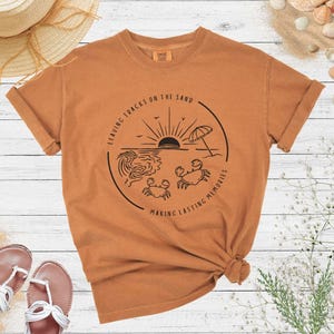 May include: A brown t-shirt with a graphic of a sunset over the ocean with two crabs and the text "Leaving tracks on the sand making lasting memories".