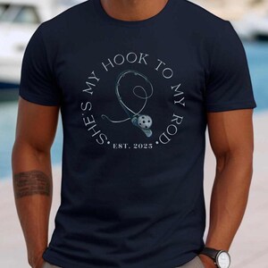 Może przedstawiać: Koszulka w kolorze granatowym z białym kołem graficznym przedstawiającym wędkę i kołowrotek z tekstem "She's my hook to my rod. Est. 2025."