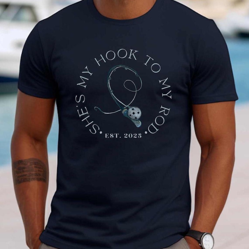 Może przedstawiać: Koszulka w kolorze granatowym z białym kołem graficznym przedstawiającym wędkę i kołowrotek z tekstem "She's my hook to my rod. Est. 2025."