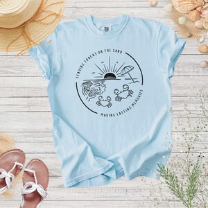 May include: Light blue t-shirt with a black graphic design of a beach scene with a sunset, waves, an umbrella, and crabs. The text on the graphic says "Leaving tracks on the sand making lasting memories".