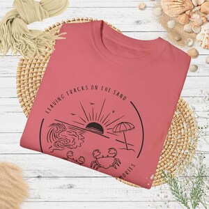 May include: A coral pink t-shirt with a black graphic design of a beach scene with a sun, waves, an umbrella, and two crabs. The text "Leaving Tracks on the Sand" is written in a circle around the graphic. The text "Memories" is written below the graphic.